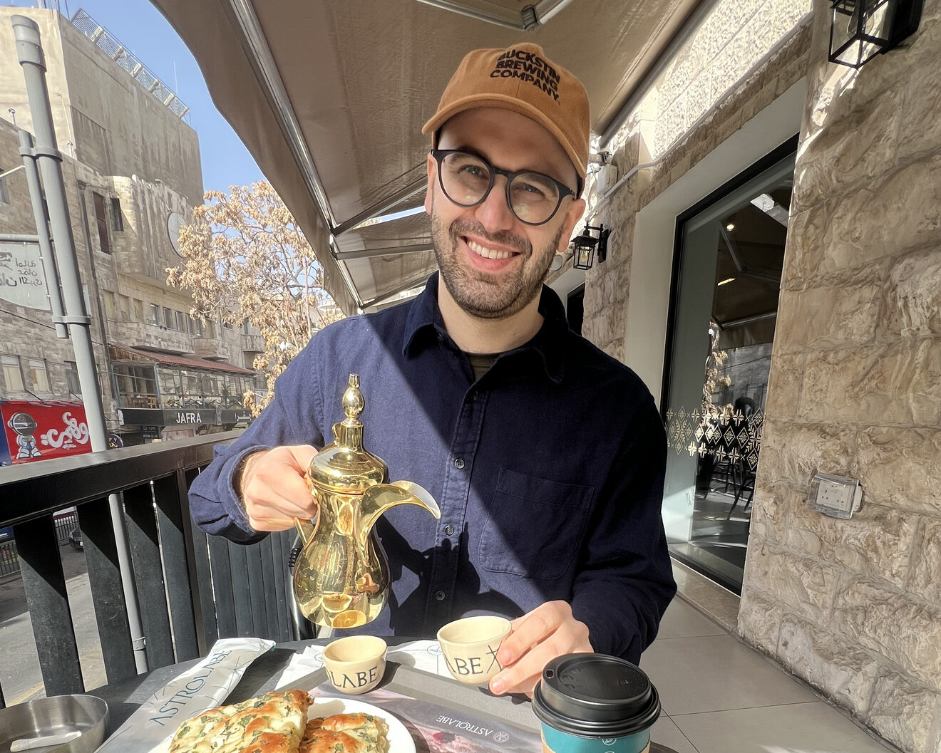 Evan, in a cap and glasses, smiling and pouring coffee from a brass dallah on a sunlit café terrace, with Amman’s stone buildings behind